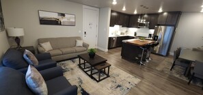 A modern living room with a sofa, coffee table, and a rug. It includes a kitchen with dark cabinets, stainless steel appliances, and a small island with a plant.