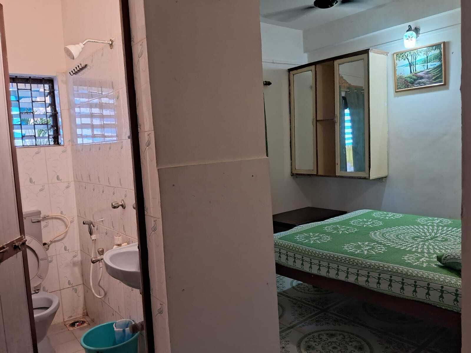 Bagno con toilette, lavandino e doccia. Si vede un letto con copriletto verde e un tappeto a motivi. Uno specchio è appeso alla parete, e una finestra con inferriate.