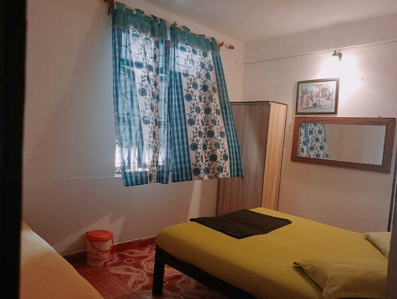 A room with a bed, a window with curtains, a mirror, and a small table with a container.