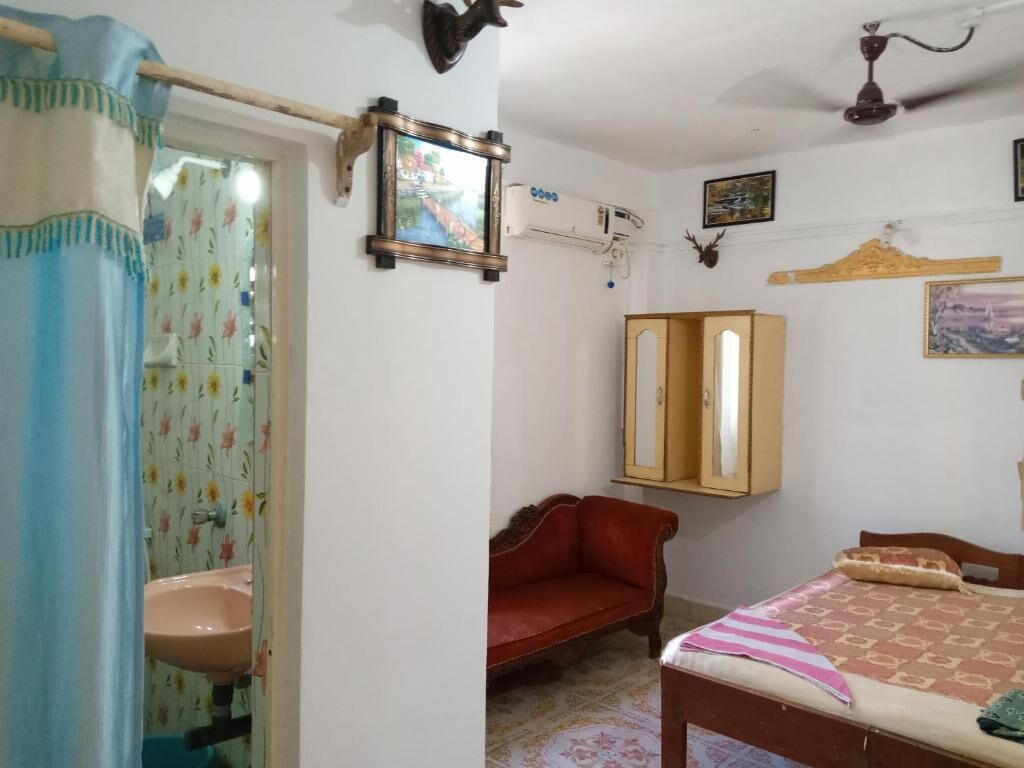 A bedroom with a bed, a wooden wardrobe, a red sofa, a wall-mounted air conditioner, and a bathroom with a sink and shower curtain.