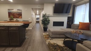 A modern kitchen with a central island, a dining area with a sofa and coffee table, and a living room with a TV and a large window.