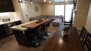 A modern kitchen with a central island, bar stools, and a dining table with chairs. There is a large window providing natural light and a view of the outdoors. - SageCreek Moab - Condo F5 - "Heated Pool - HotTub" (Moab)