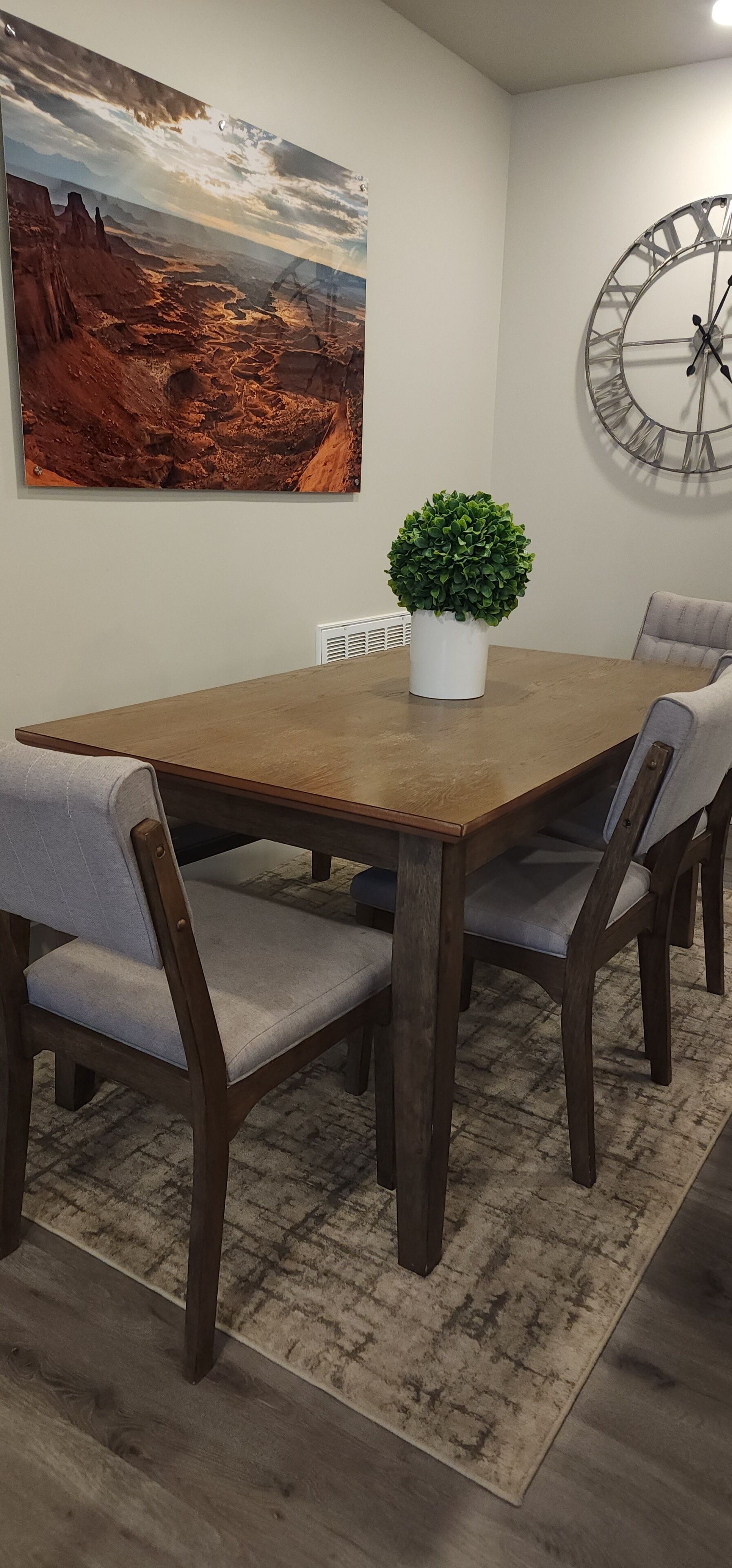 Un coin repas avec une table en bois, des chaises rembourrées, une plante en pot, une grande horloge et un tableau représentant un paysage désertique accroché au mur.
