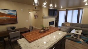 A modern living room with a wooden dining table, a gray sofa, and a TV mounted on the wall. - Specials - F1,F2,F3,F4- Sage Creek Quad 1stFloor (Moab)