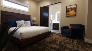 A bedroom with a large bed, a nightstand, a lamp, a chair, a plant, and a painting on the wall. - Sage Creek at Moab "Combo F10 & F11" Heated Pool (Moab)