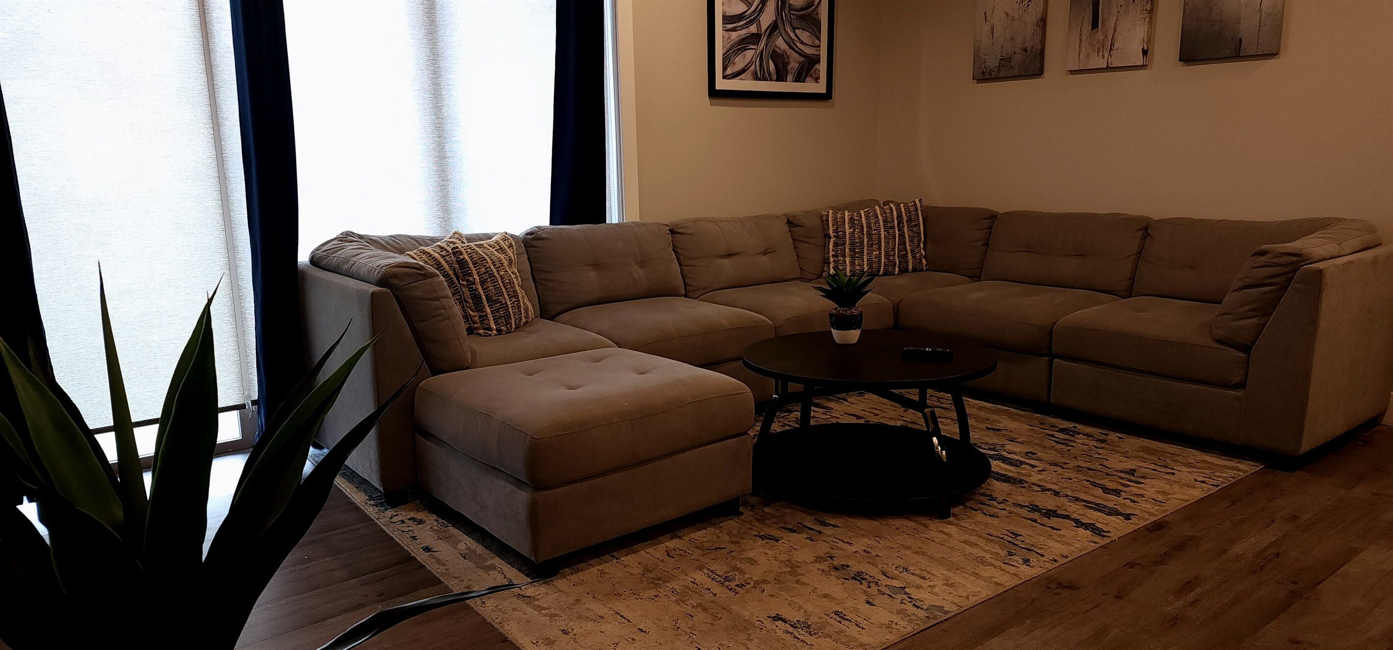 A living room with a brown sectional sofa, a small ottoman, a coffee table, and a potted plant.