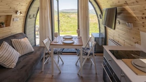 A cozy interior with a wooden dining table set for two, a sofa with patterned cushions, and a kitchen area with a stove and a flat-screen TV.