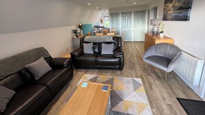A modern living room with a black leather sofa, a grey armchair, and a coffee table. A kitchen area is visible in the background with teal cabinets and a dining table. - Coombe View Cornwall Holiday Lodge Near Bude. Sea Views. Large Patio Sleeps 6. (Kilkhampton)