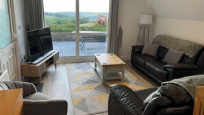 A modern living room with a flat-screen TV, a sofa, a coffee table, and a view of a green landscape through large sliding glass doors. - Coombe View Cornwall Holiday Lodge Near Bude. Sea Views. Large Patio Sleeps 6. (Kilkhampton)