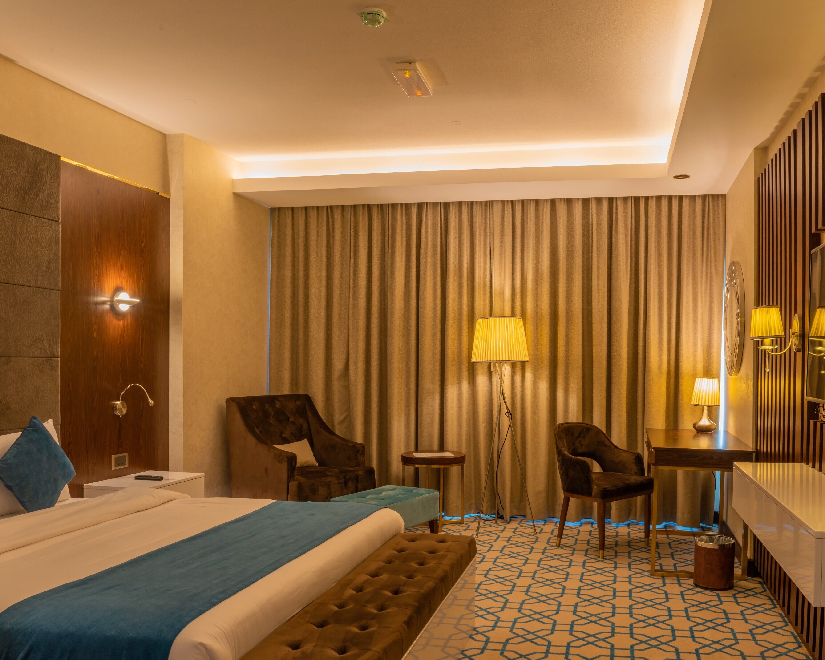 A hotel room with a bed, two chairs, a desk, a lamp, a mirror, and a curtain.