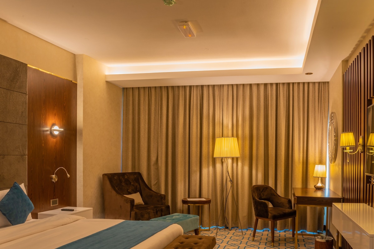 A hotel room with a bed, two chairs, a desk, a lamp, a mirror, and a curtain.
