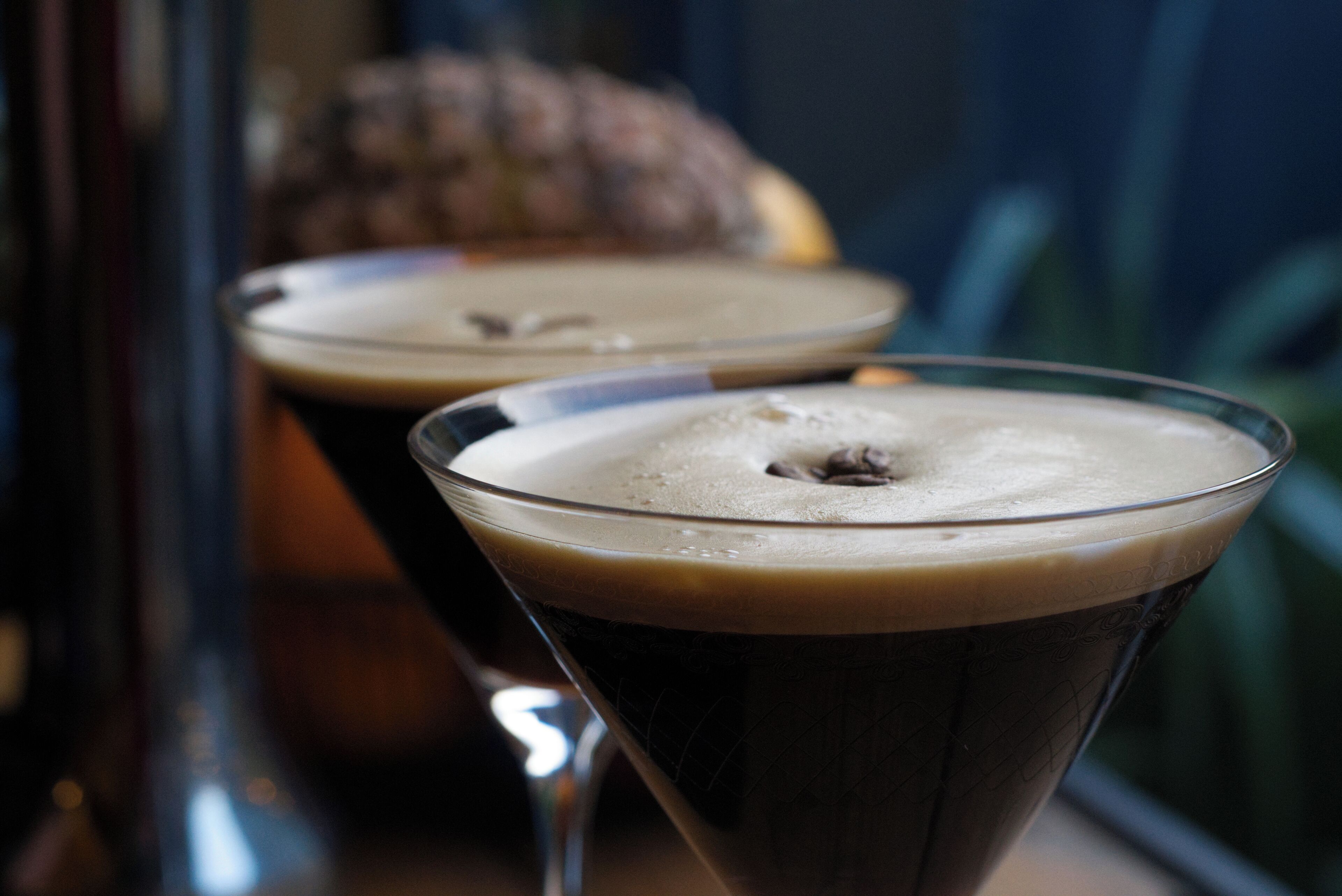 Dos vasos de martini llenos de una bebida oscura, probablemente café o té, sobre una superficie reflectante con fondo desenfocado.