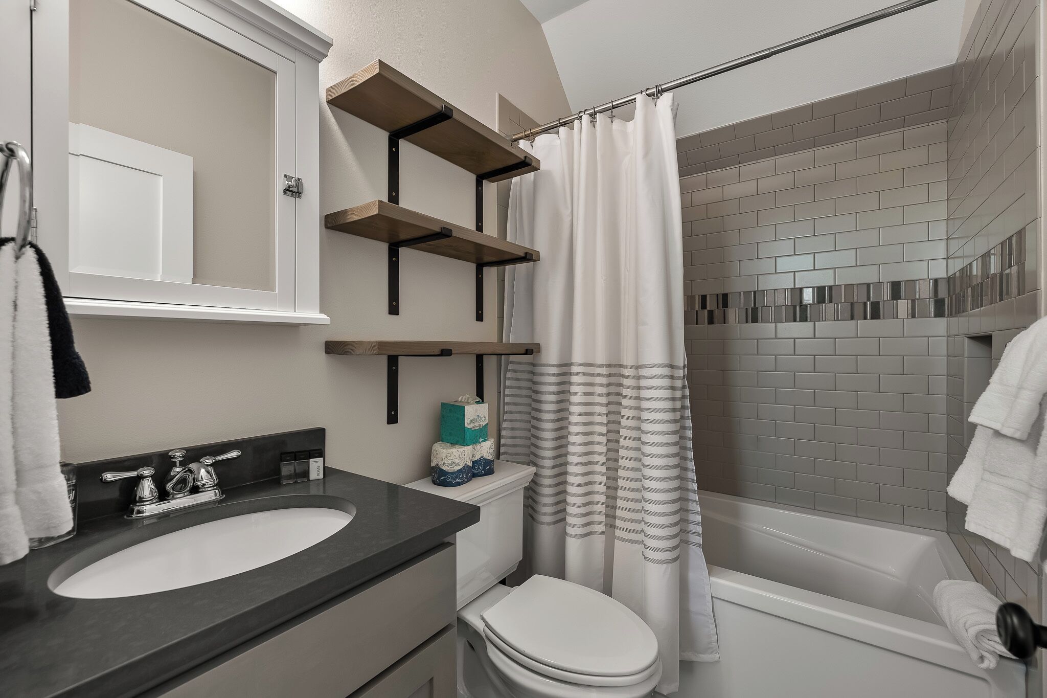A bathroom with a white toilet, a sink, a shower with a striped curtain, and a mirror with shelves above the sink.