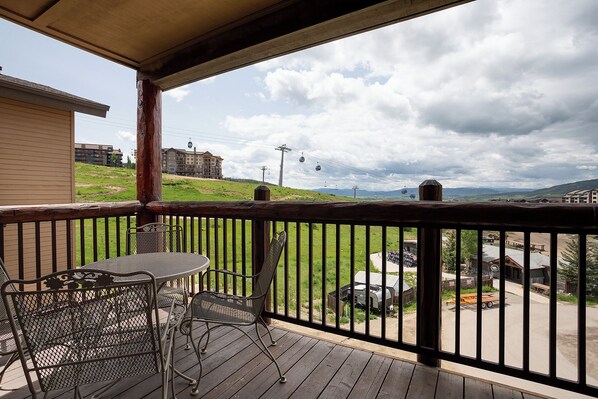 A wooden deck with metal chairs and a table, overlooking a grassy area with buildings and a distant mountain range.