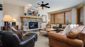 A living room with a stone fireplace, leather furniture, and wooden window blinds. - AN213 | Antlers Steamboat | Ski In/Out from Patio | Winter Shuttle (Steamboat Springs)