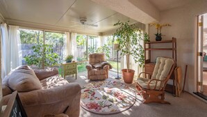A sunlit room with a ceiling fan, a round rug with floral patterns, a beige armchair, a wooden rocking chair, and a potted plant.