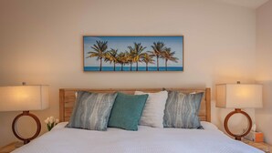 A neatly made bed with white bedding, a painting of palm trees on the wall, and two bedside lamps.