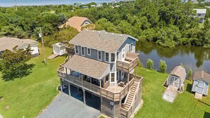 A two-story house with a large deck and a garage, surrounded by a lake and dense greenery. - Oceanside Home with Hot Tub, Nearby Beach Access, and Grills. A Retreat for All! (Salvo)