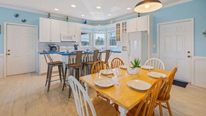 A modern kitchen with white cabinets, a central island, and a dining area with a wooden table and chairs. - Oceanside Home with Hot Tub, Nearby Beach Access, and Grills. A Retreat for All! (Salvo)
