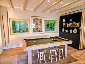 Une table de billard avec des tabourets, trois fenêtres donnant sur un espace verdoyant et une étagère noire ornée d’objets décoratifs.