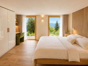 A modern bedroom with a large bed, a wooden wardrobe, a green chair, and a view of greenery and mountains through the windows.