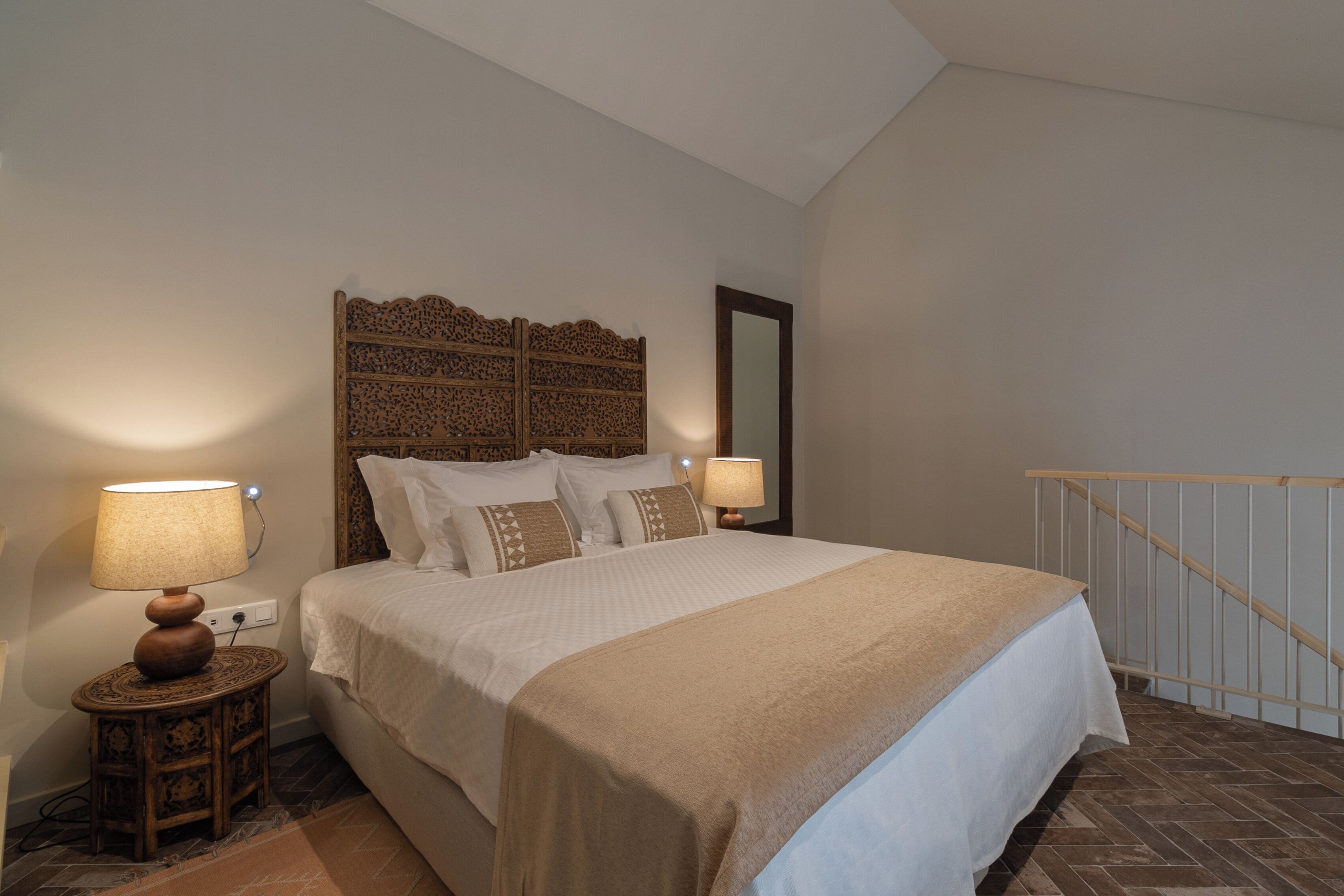 Een netjes opgemaakt bed met een houten hoofdbord, twee bedlampen en een klein houten nachtkastje.