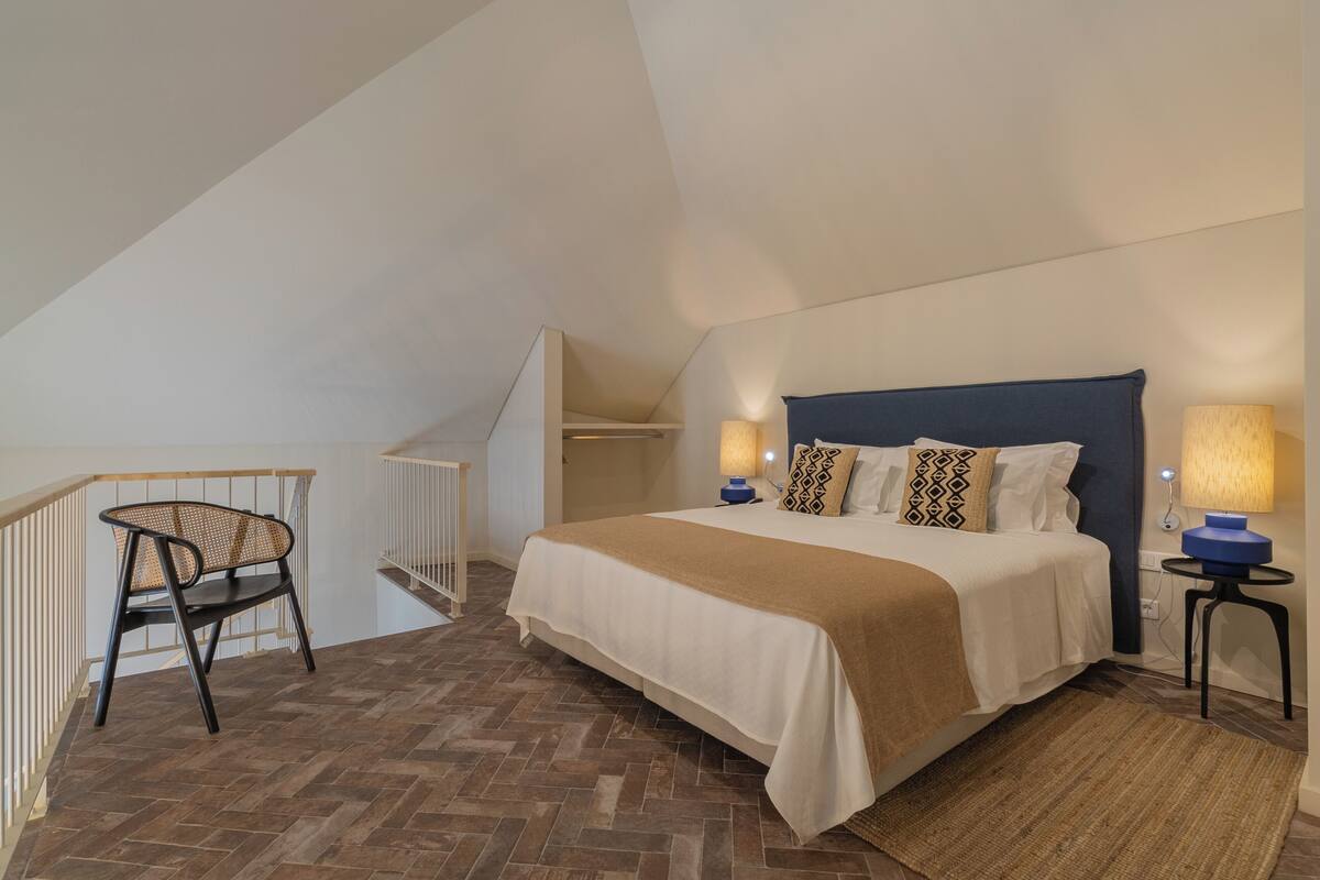 A bedroom with a bed, a chair, a lamp, and a staircase.