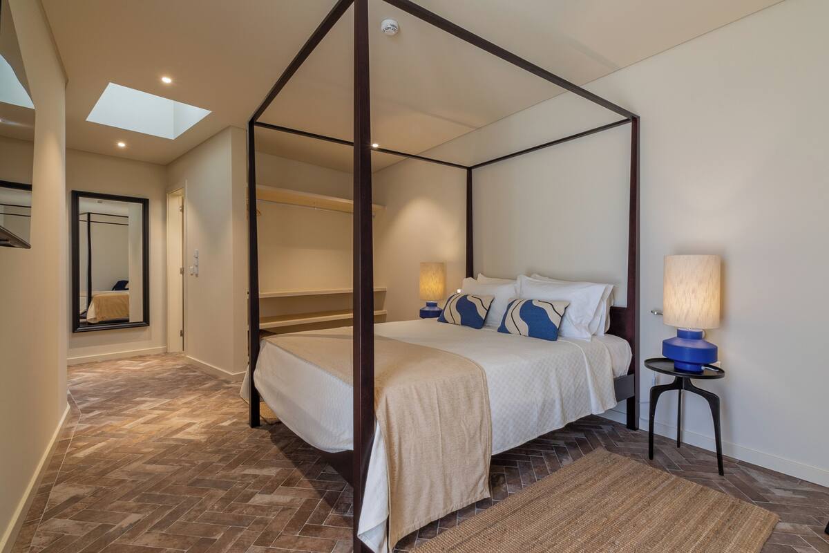 A four-poster bed with a canopy, a bedside lamp, and a view of a hallway with a mirror and a door.