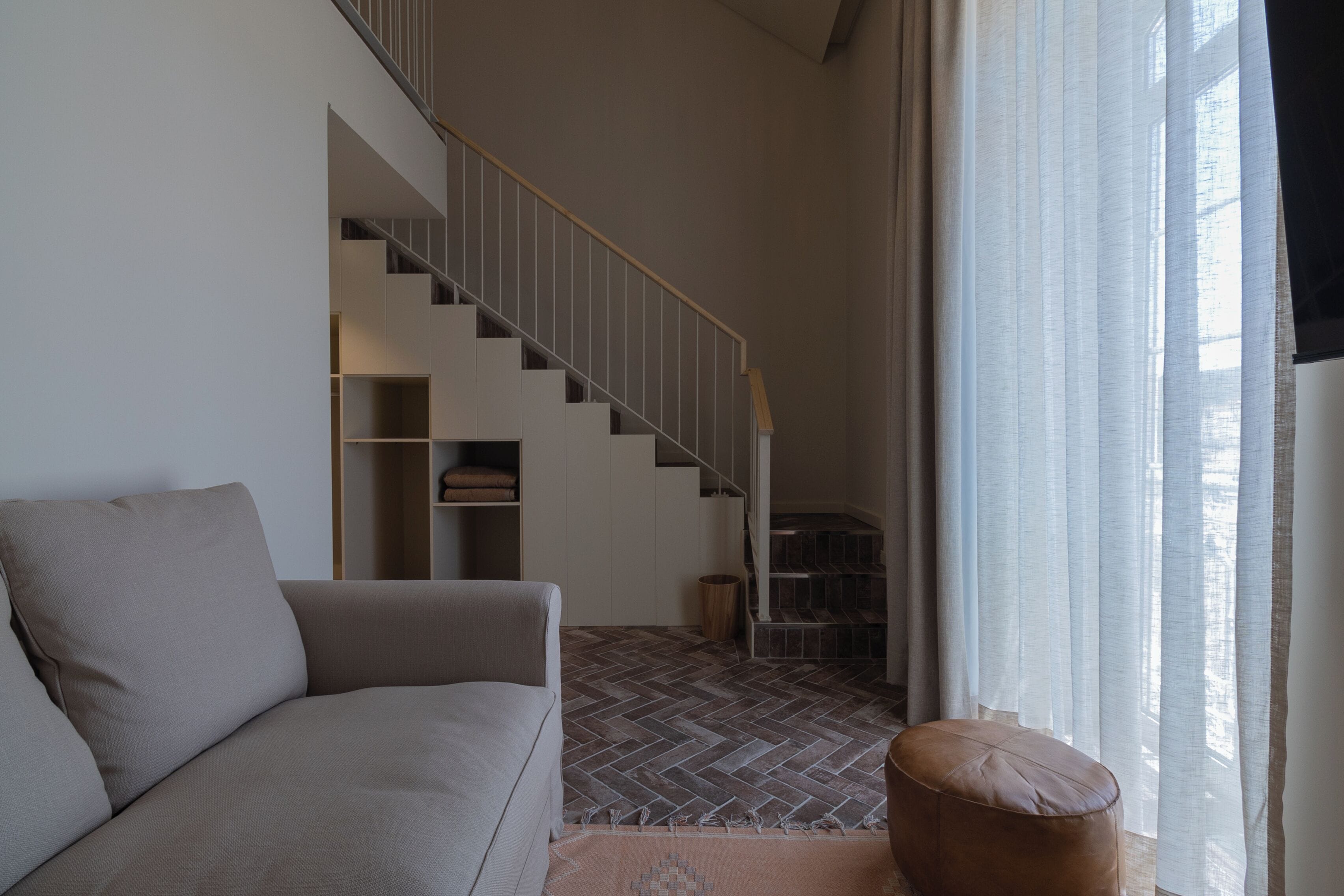 Una sala de estar moderna con sofá, un banco de madera, piso con patrón de espiga, una escalera y un gran ventanal con cortinas traslúcidas.