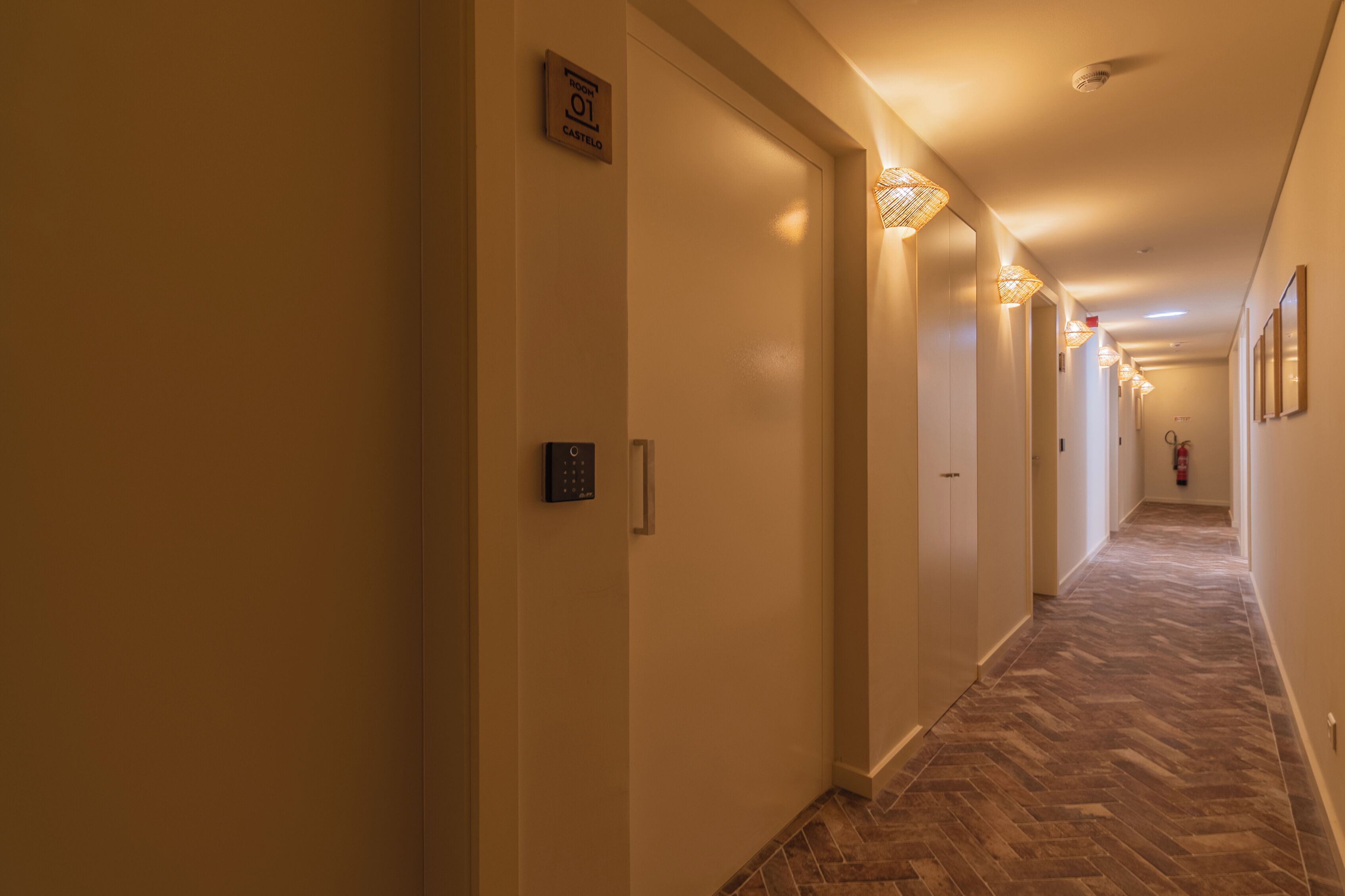 Un pasillo de hotel con piso de espiga, luminarias fijadas a la pared y una puerta con letrero indicando el número de habitación 01.