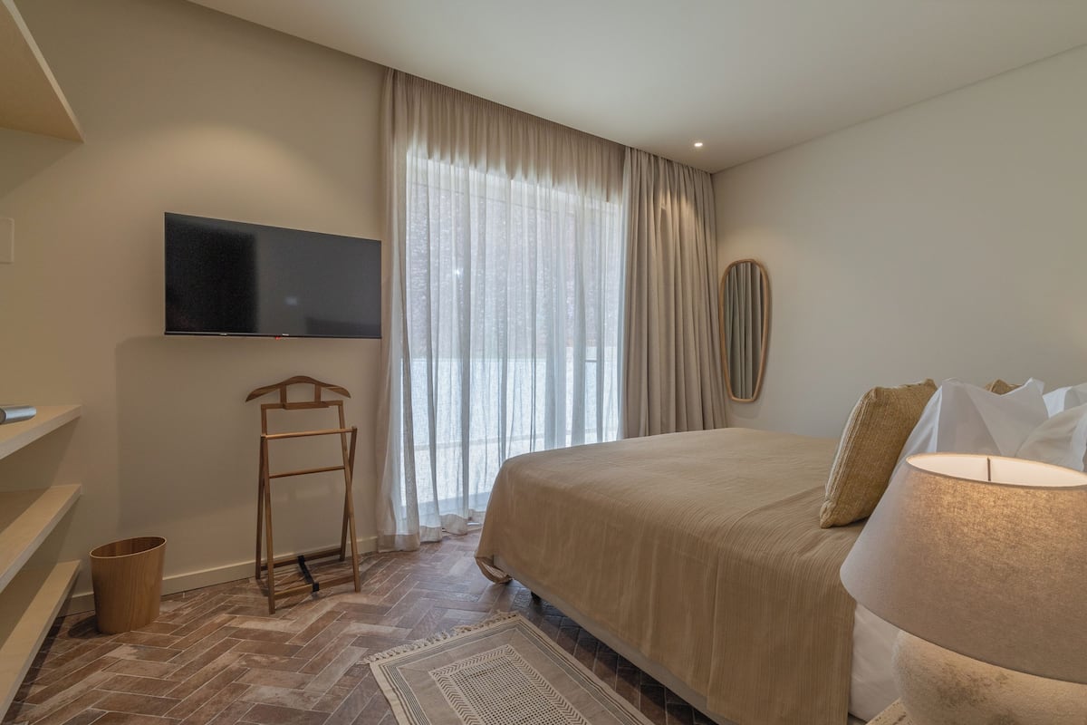 A bedroom with a bed, a TV mounted on the wall, a wooden ladder, a small trash bin, and a window with sheer curtains.