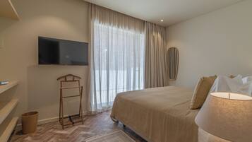 Quarto com cama, televisão na parede, escada de madeira, lixeira pequena e janela com cortinas leves.