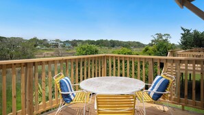 A wooden deck with a round table and two chairs, overlooking a scenic landscape. - The Marsh: Cozy Cottage with Beautiful Marsh Views, Short Walk to Beach (Oak Island)