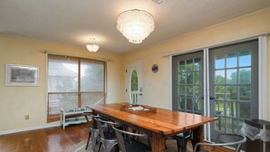 A dining area with a wooden table and metal chairs, a chandelier, and large windows with blinds. - The Marsh: Cozy Cottage with Beautiful Marsh Views, Short Walk to Beach (Oak Island)