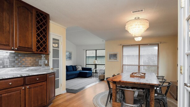 A kitchen with wooden cabinets, a marble countertop, and a chandelier. There is a dining area with a wooden table and chairs, and a living room with a blue sofa. - The Marsh: Cozy Cottage with Beautiful Marsh Views, Short Walk to Beach (Oak Island)