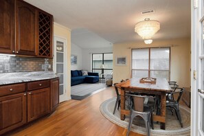 A kitchen with wooden cabinets, a marble countertop, and a chandelier. There is a dining area with a wooden table and chairs, and a living room with a blue sofa. - The Marsh: Cozy Cottage with Beautiful Marsh Views, Short Walk to Beach (Oak Island)