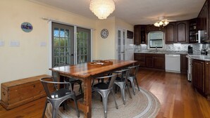 A kitchen with wooden cabinets, a dining table with chairs, a chandelier, and a clock on the wall. - The Marsh: Cozy Cottage with Beautiful Marsh Views, Short Walk to Beach (Oak Island)