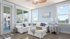 Un salón amplio con un sofá seccional blanco, una mesita auxiliar, un ventilador de techo y ventanas grandes que ofrecen vistas al exterior.