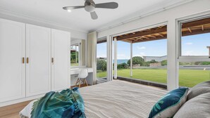 Ein Schlafzimmer mit einem großen Bett, einem Deckenventilator und Blick auf eine grüne Fläche und das Meer durch große Fenster.