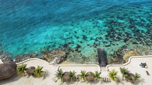 Vista aérea de un complejo turístico en la playa, con aguas turquesas cristalinas, una playa de arena y sombrillas de techo de paja.