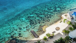 Vista aérea de un complejo turístico en la playa, con aguas turquesas cristalinas, una playa de arena y un edificio del complejo con piscina y zona de descanso.