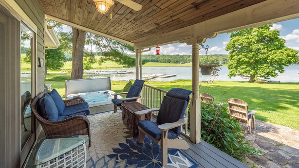 A covered outdoor area with wicker furniture, a glass table, and a view of a lake and greenery. - Dog Friendly Cottage w/ Dock & Shuffleboard (Swanton)