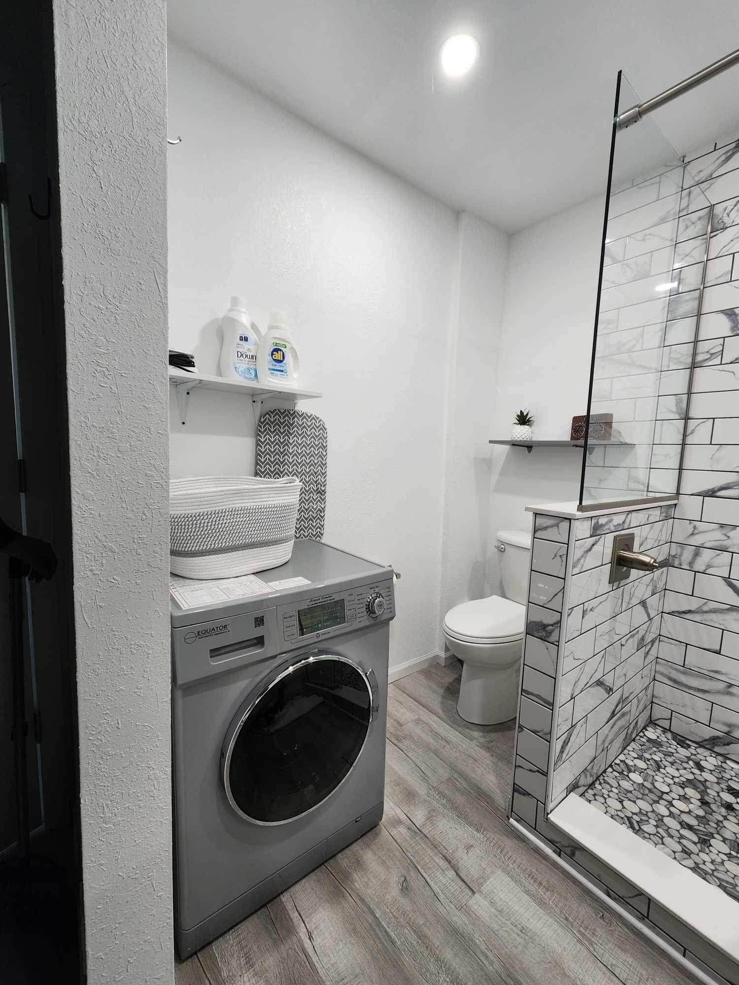 Une salle de bain moderne équipée d’un lave-linge intégré, de toilettes et d’une douche à parois de verre.