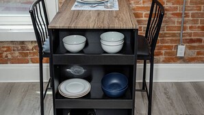 A dining table with a wooden top, black chairs, and a black shelving unit below holding bowls, plates, and a coffee maker.