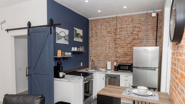 A compact kitchen with a blue door, white cabinets, a stainless steel refrigerator, and a brick wall.