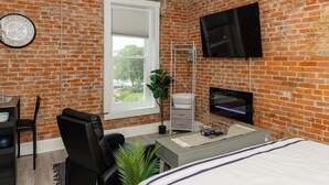 A modern bedroom with a brick wall, a flat-screen TV, a desk with a chair, a bed, and a window with a view.