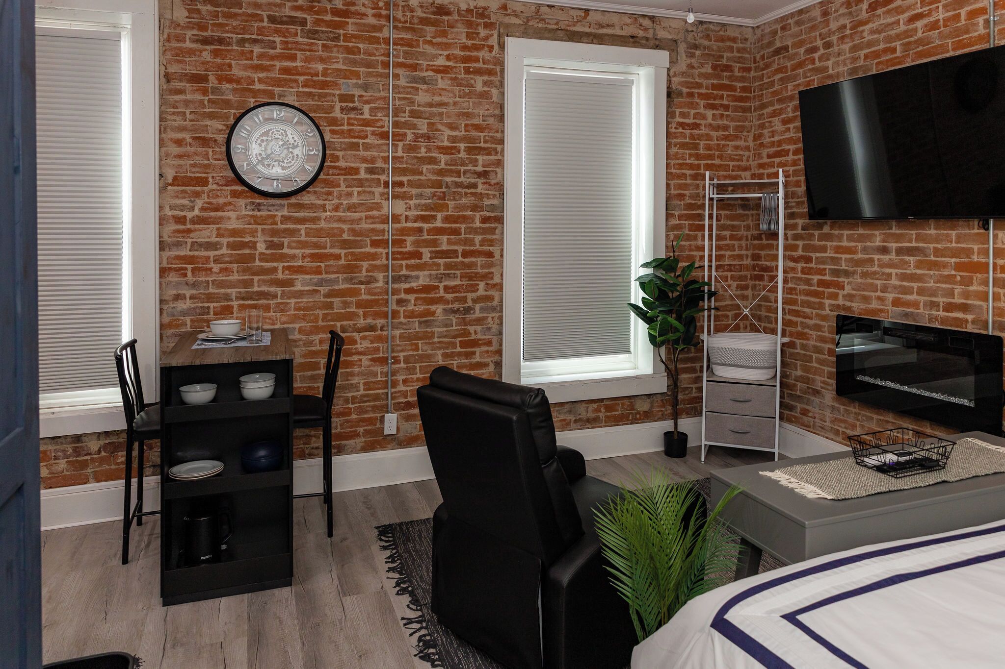 Un dormitorio moderno con pared de ladrillo, televisor de pantalla plana, cama, escritorio con silla y ventana con persianas.
