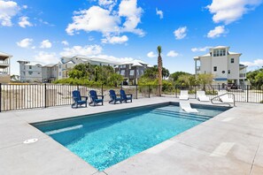 A rectangular swimming pool with clear blue water, surrounded by a black metal fence. There are several lounge chairs, some blue and some white, along the poolside. The pool area is adjacent to a building with a white exterior and a flat roof.