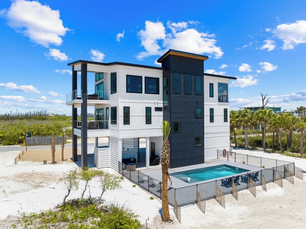 A modern multi-story building with a pool, surrounded by a sandy beach and palm trees.
