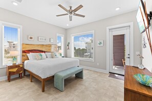 A bedroom with a bed, bench, bedside tables, ceiling fan, windows, and a door.
